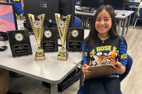 A girl in a CE Rose shirt sits next to several trophies