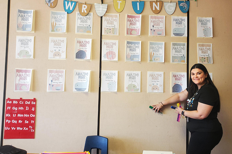 A woman in a black shirt points to her classroom wall