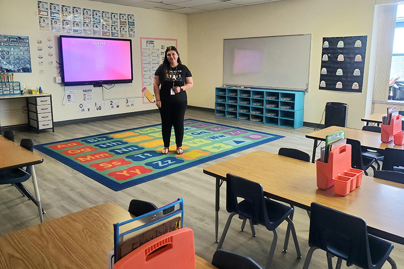 A woman in a black shirt and pants stands on a rug in her classroom