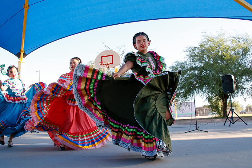 Three girls in colorful folklorico dresses dance on the basketball court outside