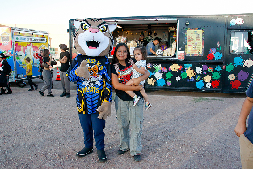 A mom holds her young daughter, standing next to the CE Rose tiger mascot, in front of a food truck