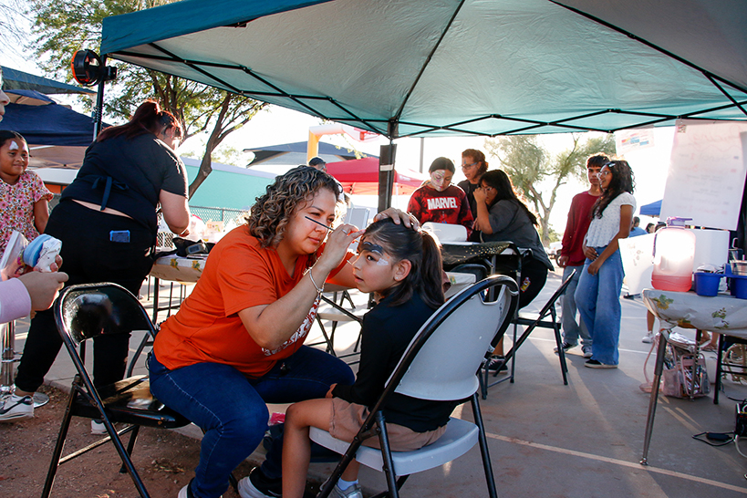 A girl sits in a chair as a woman paints a bat on her forehead
