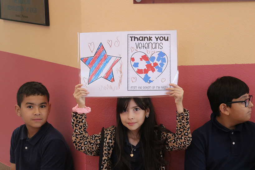 A girl holds up a coloring page with the words Thank You Veterans and colored images of a star and heart
