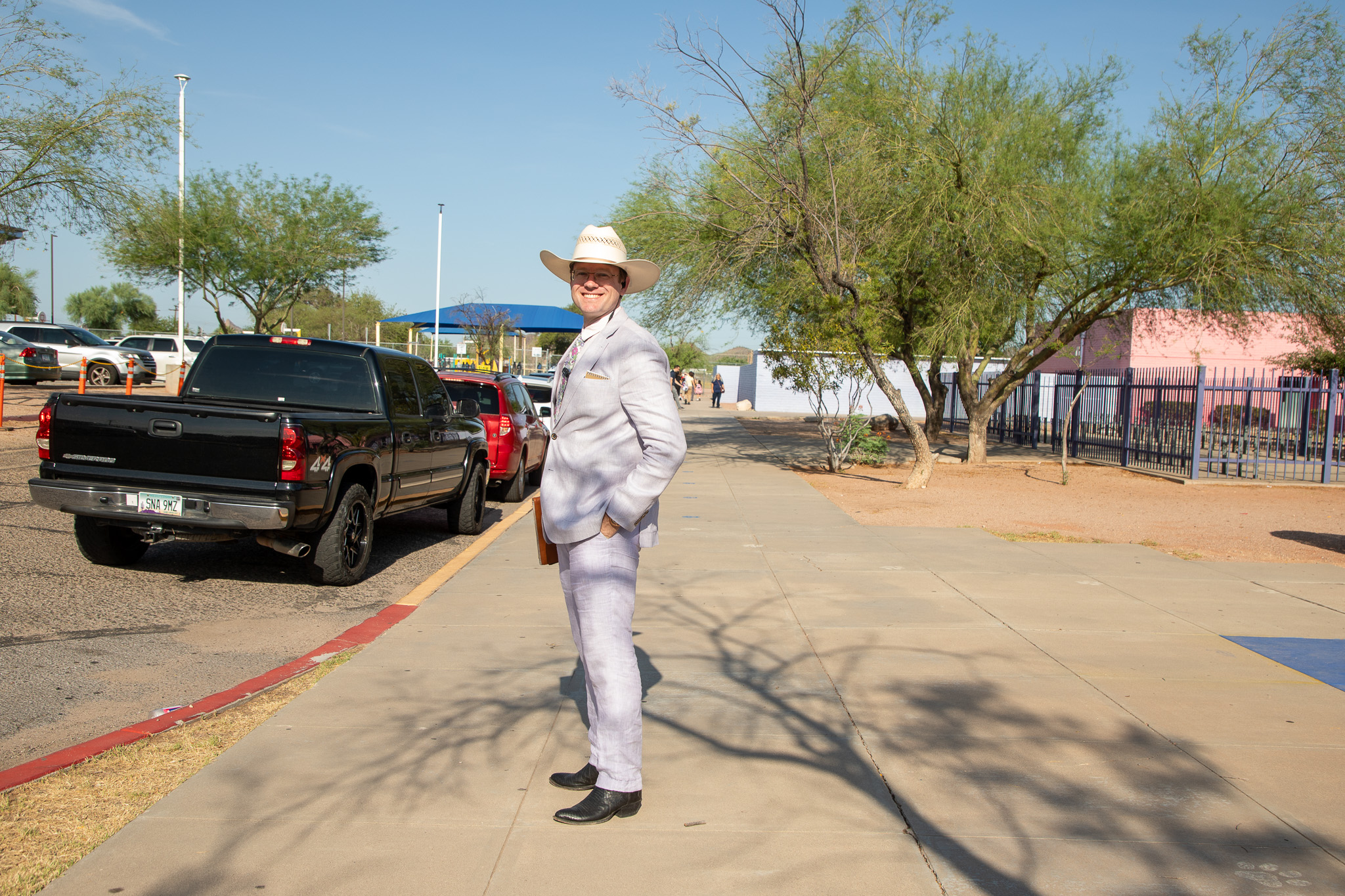 A man in a white suit and cowboy hat smiles on the curb in front of the school